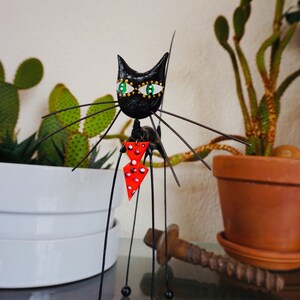 Puede incluir: Figurilla de gato negro con ojos verdes y una pajarita roja con lunares blancos. El gato est&aacute; hecho de alambre y tiene un cuerpo largo y delgado. La figurilla est&aacute; de pie sobre una mesa con una planta en maceta en el fondo.