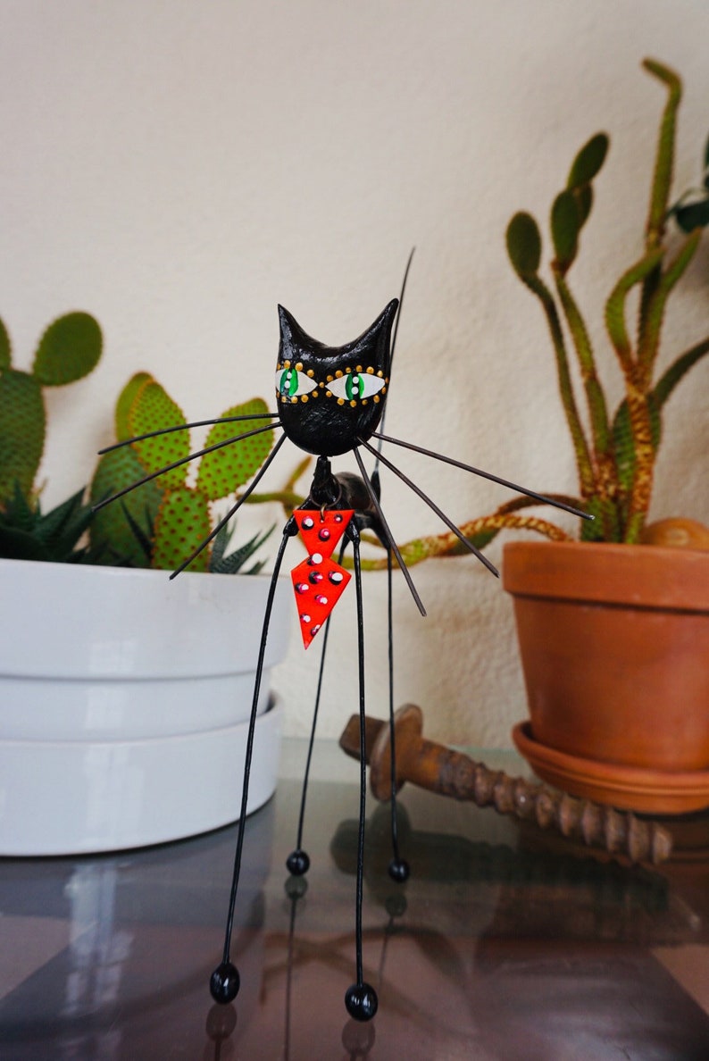 Puede incluir: Figurilla de gato negro con ojos verdes y una pajarita roja con lunares blancos. El gato est&aacute; hecho de alambre y tiene un cuerpo largo y delgado. La figurilla est&aacute; de pie sobre una mesa con una planta en maceta en el fondo.