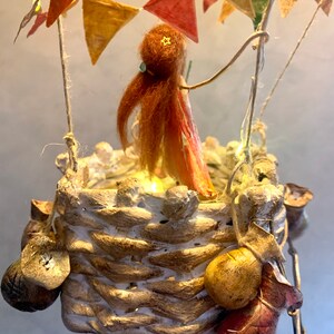 Może przedstawiać: Kapryśna, ręcznie robiona rzeźba balonu na ogrzane powietrze. Kosz wykonany jest z teksturowanego, jasnego materiału, z dekoracyjnymi elementami wiszącymi. W środku stoi mała postać z rudymi włosami i gwiazdą na głowie. Kolorowe chorągiewki i lampki choinkowe zdobią g&oacute;rę.