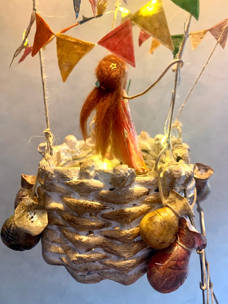 Może przedstawiać: Kapryśna, ręcznie robiona rzeźba balonu na ogrzane powietrze. Kosz wykonany jest z teksturowanego, jasnego materiału, z dekoracyjnymi elementami wiszącymi. W środku stoi mała postać z rudymi włosami i gwiazdą na głowie. Kolorowe chorągiewki i lampki choinkowe zdobią g&oacute;rę.