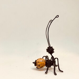 Puede incluir: Escultura de alambre hecha a mano de una hormiga que lleva una bola moteada. La hormiga est&aacute; hecha de alambre negro, con un cuerpo enrollado y largas antenas. La bola es de color marr&oacute;n claro con motas marrones oscuras.
