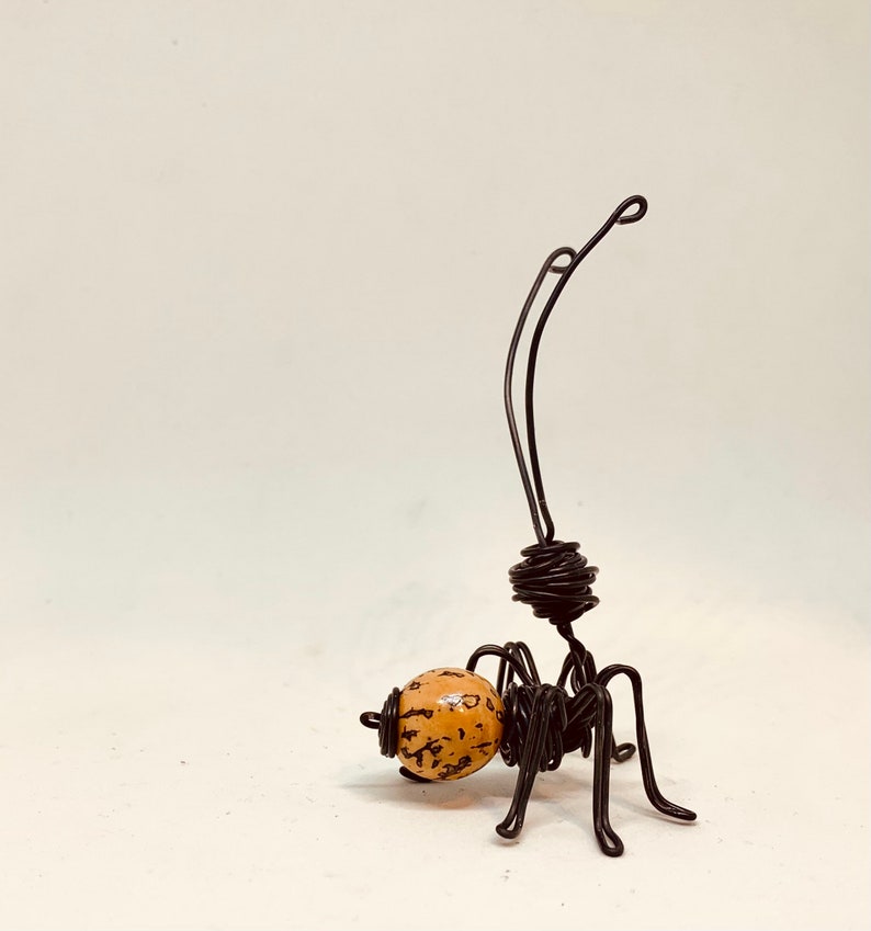 Puede incluir: Escultura de alambre hecha a mano de una hormiga que lleva una bola moteada. La hormiga est&aacute; hecha de alambre negro, con un cuerpo enrollado y largas antenas. La bola es de color marr&oacute;n claro con motas marrones oscuras.
