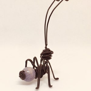 Puede incluir: Una escultura de alambre hecha a mano de una hormiga. La hormiga presenta un cuerpo de alambre marr&oacute;n oscuro, seis patas y dos largas antenas. La cabeza es una cuenta de color p&uacute;rpura claro. Una obra de arte &uacute;nica.