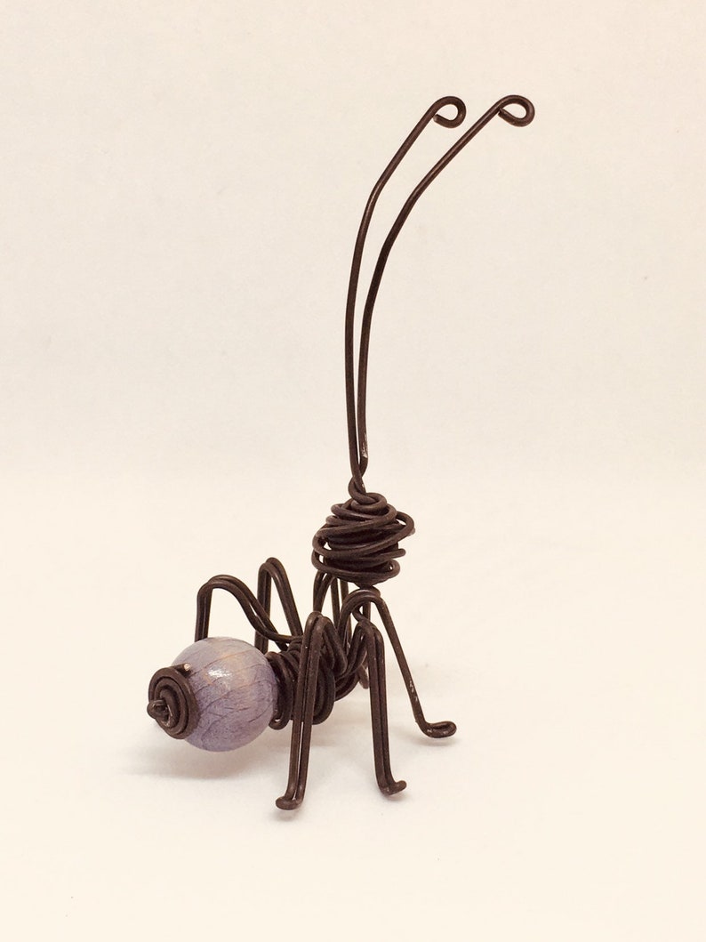 Puede incluir: Una escultura de alambre hecha a mano de una hormiga. La hormiga presenta un cuerpo de alambre marr&oacute;n oscuro, seis patas y dos largas antenas. La cabeza es una cuenta de color p&uacute;rpura claro. Una obra de arte &uacute;nica.