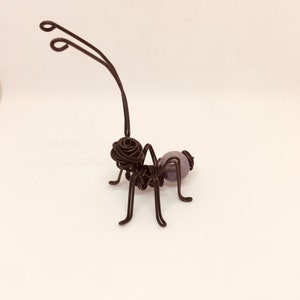 Puede incluir: Una escultura artesanal de alambre de una hormiga. La hormiga est&aacute; hecha de alambre marr&oacute;n oscuro con un cuerpo redondeado y seis patas. El cuerpo de la hormiga presenta una cuenta esf&eacute;rica p&uacute;rpura. Las antenas se curvan hacia arriba.