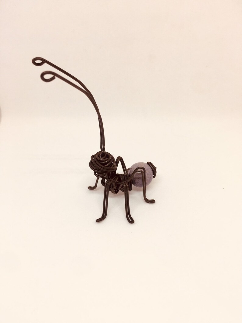 Puede incluir: Una escultura artesanal de alambre de una hormiga. La hormiga est&aacute; hecha de alambre marr&oacute;n oscuro con un cuerpo redondeado y seis patas. El cuerpo de la hormiga presenta una cuenta esf&eacute;rica p&uacute;rpura. Las antenas se curvan hacia arriba.