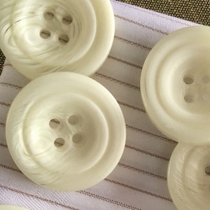 Puede incluir: Diez botones de plástico blanco con cuatro agujeros cada uno. Los botones tienen una superficie ligeramente texturizada y un borde ligeramente elevado.