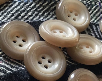 May include: A collection of 12 beige and white buttons with four holes each. The buttons are arranged on a beige and black patterned fabric and a beige knitted fabric.