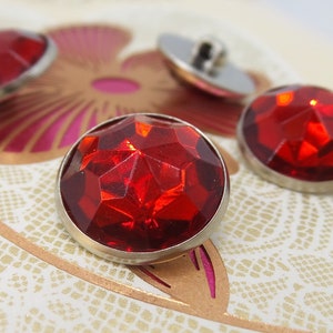 Puede incluir: Cuatro botones de cristal rojo facetados con monturas de metal plateado. Los botones están dispuestos sobre un fondo blanco con un diseño floral.
