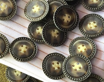May include: A collection of round, brown and cream colored buttons with a decorative metal rim. The buttons have four holes and are arranged in a random pattern.