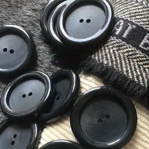 May include: A collection of black plastic buttons with two holes each. The buttons are arranged on a white and black patterned fabric.