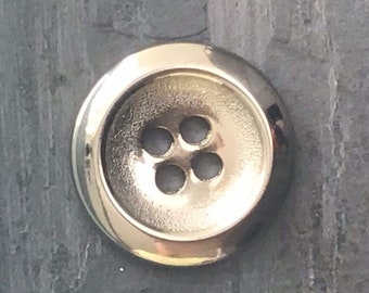 May include: A shiny, gold-colored button with four holes. The button is round with a slightly raised center. The button is approximately 0.75 inches in diameter. The button is on a dark gray surface.