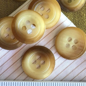 May include: Eight brown, four-hole buttons with a slightly raised edge and a smooth, shiny finish. The buttons are arranged on a white fabric with brown stripes and a ruler measuring 1 inch.