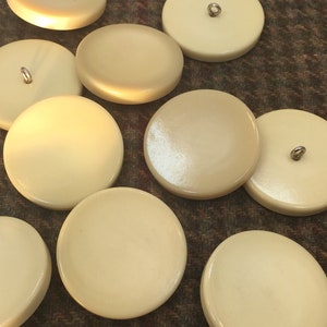 May include: A collection of ten off-white round buttons with a hole in the center. The buttons are smooth and have a slight sheen.