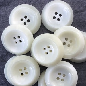 May include: A group of eight white plastic buttons with four holes each. The buttons are arranged in a circle on a gray surface.