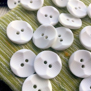 May include: A collection of white, two-hole, round buttons with a textured surface. The buttons are arranged on a green fabric background.