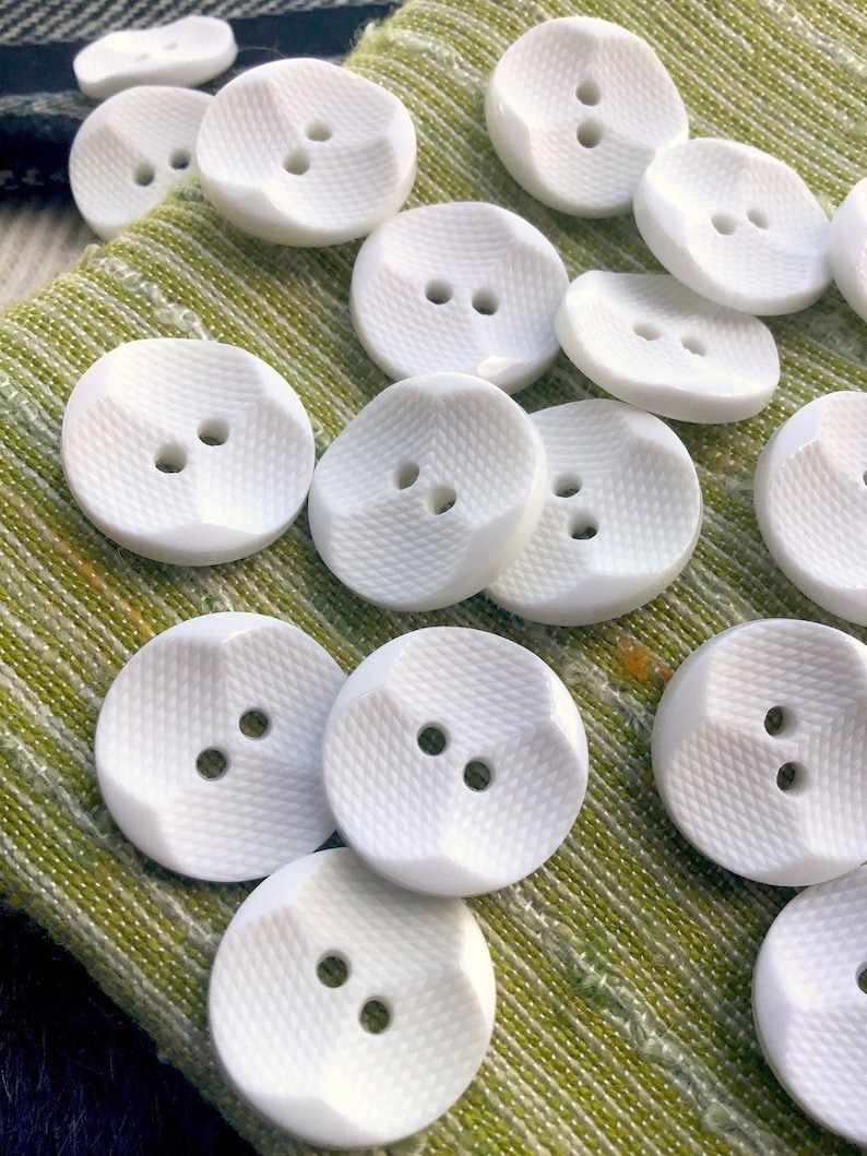 May include: A collection of white, two-hole, round buttons with a textured surface. The buttons are arranged on a green fabric background.