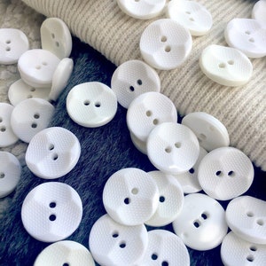 May include: A collection of white plastic buttons with two holes each. The buttons have a textured surface and are arranged in a random pattern.