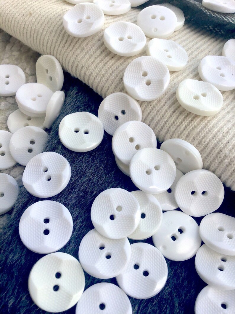 May include: A collection of white plastic buttons with two holes each. The buttons have a textured surface and are arranged in a random pattern.