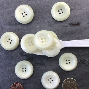 May include: A collection of eight off-white plastic buttons with four holes each. The buttons are arranged on a gray surface with a spoon and a US coin.