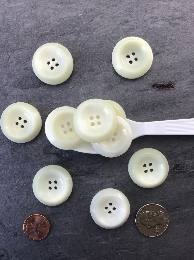 May include: A collection of eight off-white plastic buttons with four holes each. The buttons are arranged on a gray surface with a spoon and a US coin.