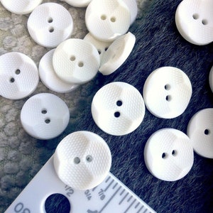 May include: A collection of white plastic buttons with two holes each. The buttons have a textured surface and are approximately 1 inch in diameter.