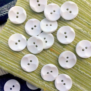 May include: A collection of white plastic buttons with two holes each. The buttons have a textured surface and are arranged on a green fabric background.