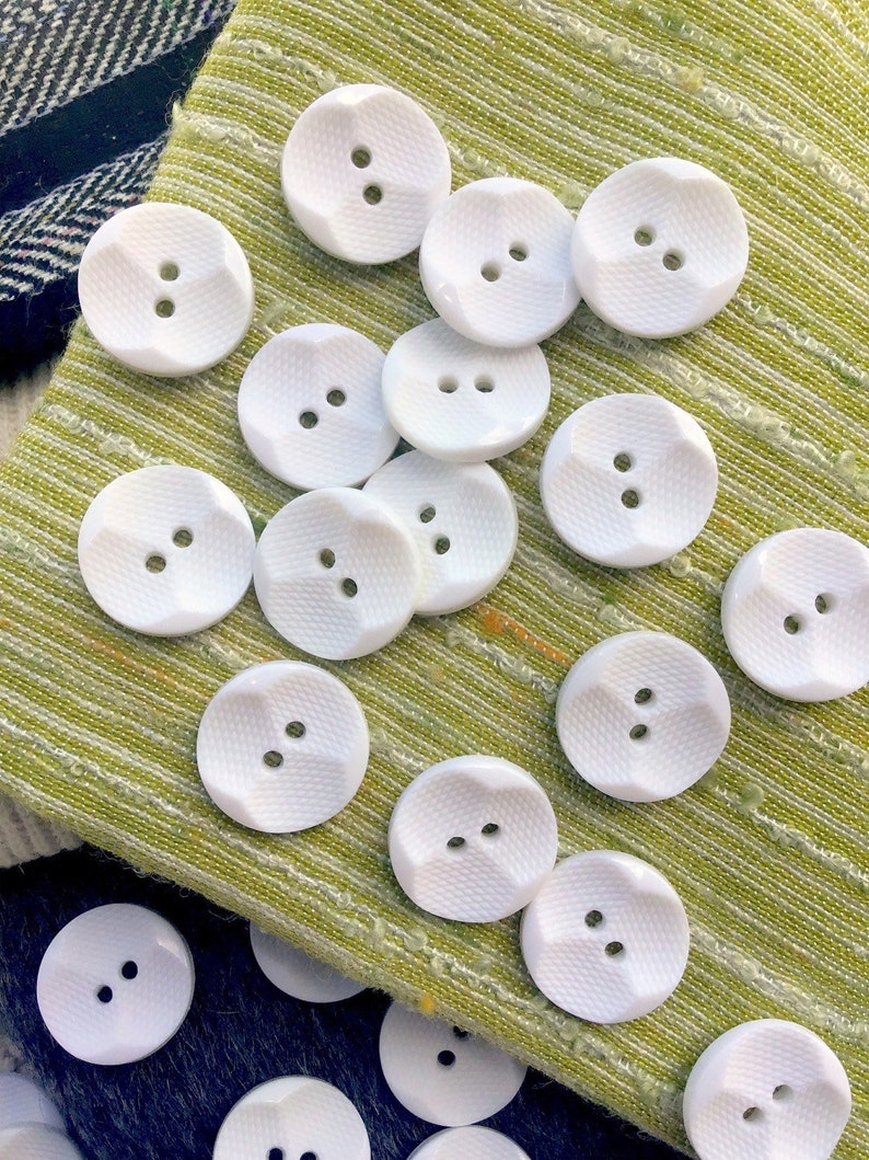 May include: A collection of white plastic buttons with two holes each. The buttons have a textured surface and are arranged on a green fabric background.