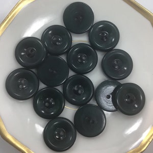 Puede incluir: Una colección de 13 botones de color verde oscuro con dos agujeros cada uno. Los botones están dispuestos en círculo sobre un plato blanco con un borde dorado.