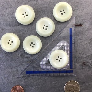 May include: Six off-white plastic buttons with four holes each. The buttons are arranged in a semi-circle with a ruler and two coins in the background.