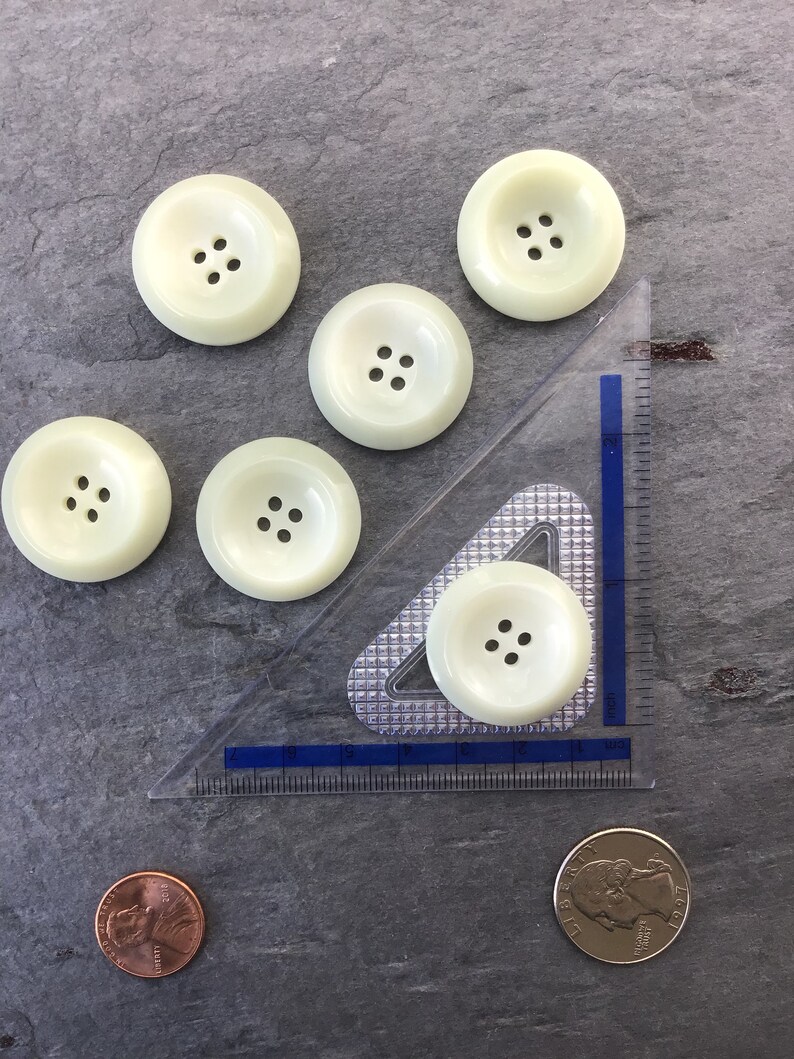 May include: Six off-white plastic buttons with four holes each. The buttons are arranged in a semi-circle with a ruler and two coins in the background.