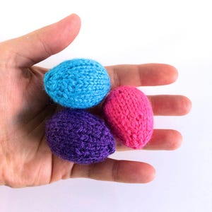 May include: Three small, egg-shaped objects crafted from knitted yarn in bright solid colours. One egg is light blue, one is deep purple, and one is vibrant pink. Each piece features a textured, looped knit pattern consistent with hand-knitted items.