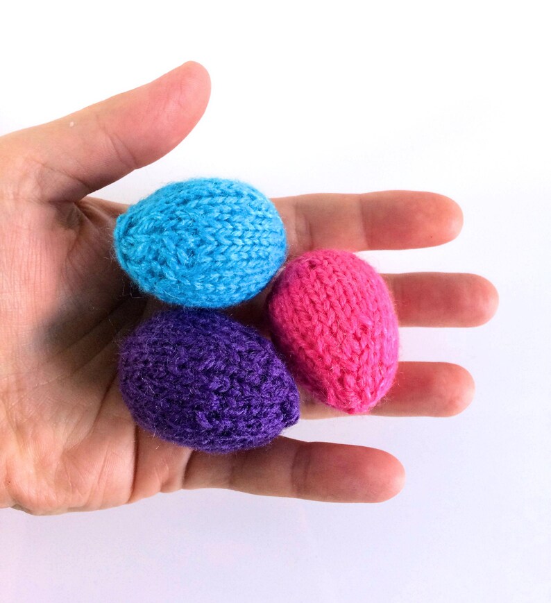 May include: Three small, egg-shaped objects crafted from knitted yarn in bright solid colours. One egg is light blue, one is deep purple, and one is vibrant pink. Each piece features a textured, looped knit pattern consistent with hand-knitted items.