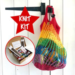 May include: A rainbow-coloured knit market bag with a mesh design hanging from a black hook, accompanied by an open cardboard box containing knitting needles, yarn skeins, and instructions labelled KNIT KIT in a red star graphic.
