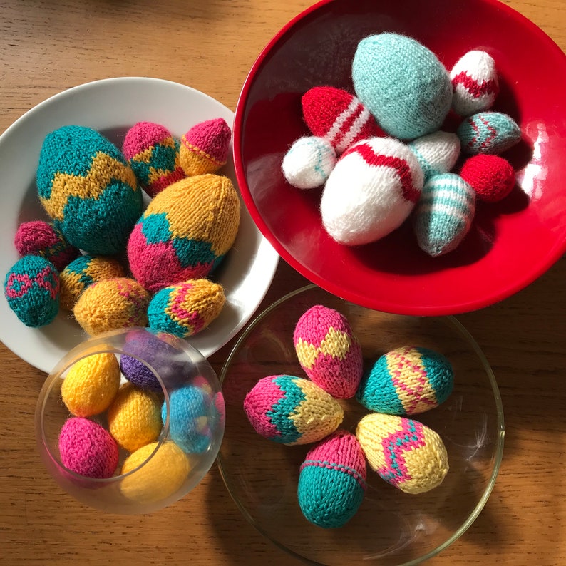 May include: A collection of knitted egg-shaped ornaments in various colours and patterns, displayed in a white plate, a red bowl, a clear glass bowl, and on a clear glass plate. The eggs feature stripes, zigzags, and solid colours.