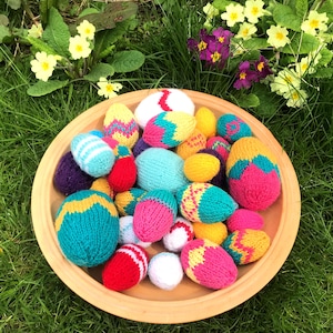 May include: A collection of small, hand-knitted egg-shaped decorations in a variety of bright colours including pink, yellow, turquoise, red, purple, and white, featuring striped and zigzag patterns, all contained within a round, light brown bowl.
