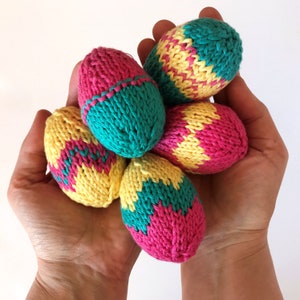 May include: Five egg-shaped objects crafted from knitted yarn in vibrant shades of pink, yellow, and teal. Each piece features a distinct colour-blocked or chevron-style pattern, showcasing the textured loops of the knit construction throughout.