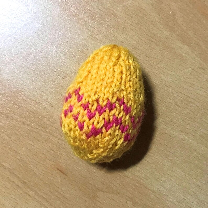May include: A small, egg-shaped object crafted from yellow yarn with a knitted texture. The surface features a repeating zigzag pattern in bright pink yarn, creating a distinct contrast against the yellow base of this soft, textile-based item.