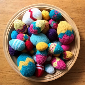 May include: A collection of small, hand-knitted egg-shaped ornaments in a round wooden bowl. The knitted eggs feature various colourful patterns, including zigzags, stripes, and solid colours in shades of yellow, blue, pink, red, white, and purple.