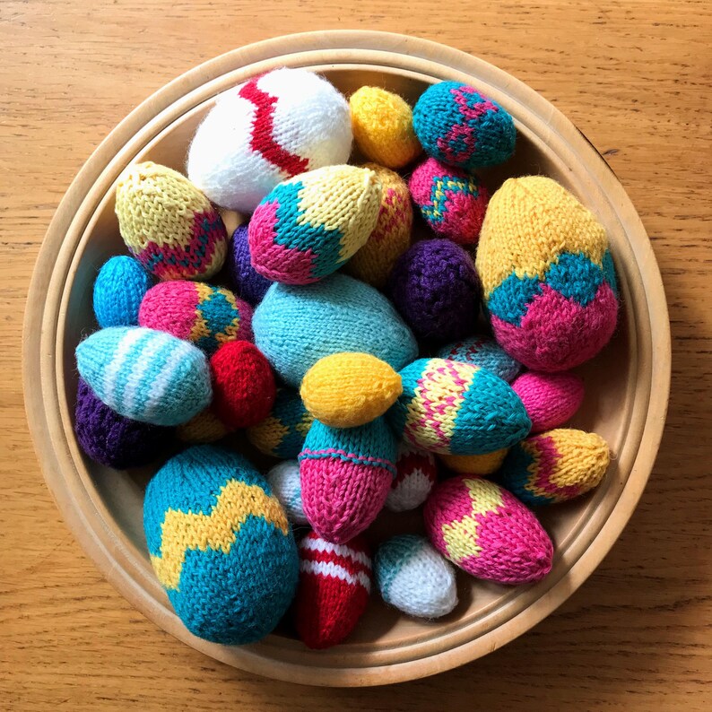 May include: A collection of small, hand-knitted egg-shaped ornaments in a round wooden bowl. The knitted eggs feature various colourful patterns, including zigzags, stripes, and solid colours in shades of yellow, blue, pink, red, white, and purple.