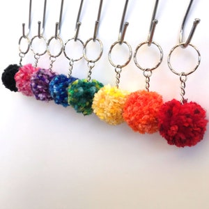 May include: A set of 9 colourful pom-pom keyrings hanging from silver key rings. The pom-poms are black, pink, purple, blue, green, yellow, orange, and red.