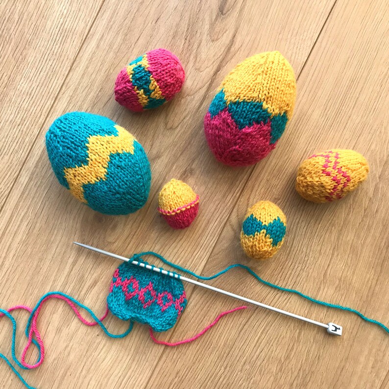 May include: A collection of six hand-knitted egg-shaped ornaments in bright yellow, turquoise, and pink yarn, featuring zigzag and diamond patterns. A single silver knitting needle holds a partially completed knitted piece with matching yarn.