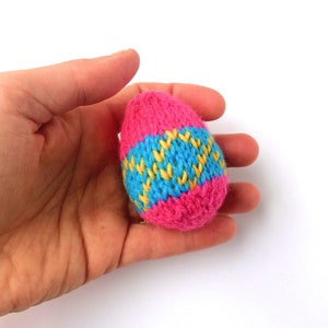 May include: A small, egg-shaped object crafted from knitted yarn. The item features vibrant horizontal bands of hot pink at the top and bottom, with a central section displaying a repeating zigzag pattern in bright yellow and light blue yarn.