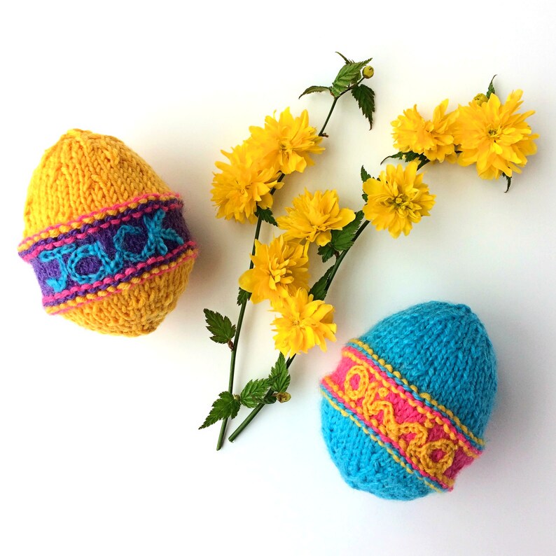 May include: Two hand-knitted egg-shaped decorations. One is yellow with a purple band featuring the name Jack in blue. The other is blue with a pink band featuring the name Olivia in yellow. A sprig of yellow flowers is positioned between them.