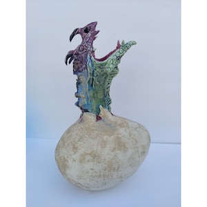 May include: A ceramic sculpture of a dragon emerging from a cracked egg. The dragon is multi-coloured with shades of green, purple, and blue. The egg is a light beige colour.