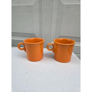 Könnte beinhalten: Zwei leuchtend orangefarbene Keramikbecher mit glatter, glänzender Oberfläche, zylindrischem Körper und einem kleinen, kreisförmigen Henkel an der Seite. Jeder Becher hat ein dezentes, erhabenes Ringdetail am oberen Rand.