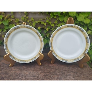 May include: Two white ceramic plates with a gold and black geometric pattern around the rim. The plates are on gold metal stands.