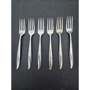 May include: Six silver forks with a floral design on the handles are arranged on a dark surface. The forks have four tines each and a curved handle with a decorative floral pattern near the base.