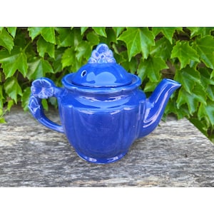 May include: A cobalt blue ceramic teapot with a lid and a curved spout. The handle and lid are adorned with floral details. The teapot is sitting on a weathered wooden surface, with green foliage in the background.