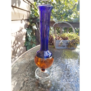Puede incluir: Un jarrón de vidrio azul y ámbar con una base redonda y un cuello alto y delgado. El jarrón está sobre una mesa de vidrio.